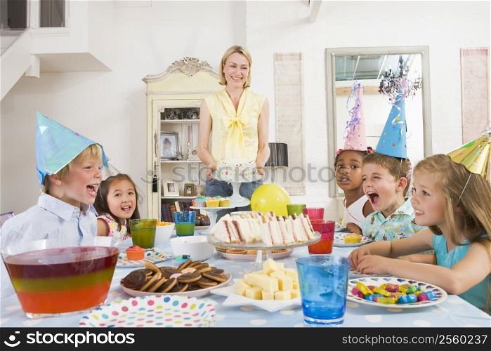 Young children at party sitting at table with mother carrying cake and smiling