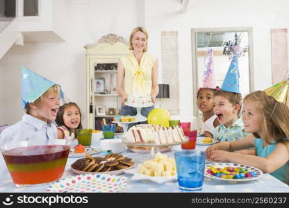 Young children at party sitting at table with mother carrying cake and smiling