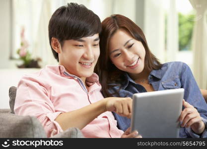 Young Chinese Couple Using Digital Tablet Whilst Relaxing On Sofa At Home