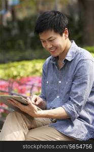 Young Chinese Man Using Digital Tablet Whilst Relaxing On Park Bench