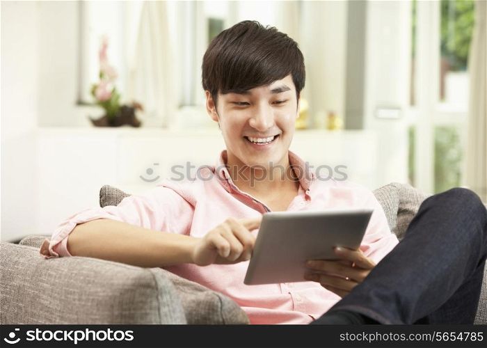 Young Chinese Man Using Digital Tablet Whilst Relaxing On Sofa At Home