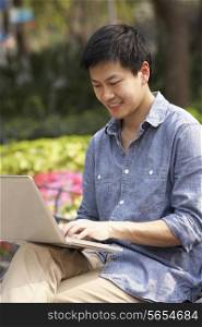 Young Chinese Man Using Laptop Whilst Relaxing On Park Bench