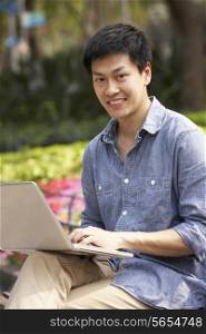Young Chinese Man Using Laptop Whilst Relaxing On Park Bench