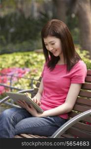 Young Chinese Woman Using Digital Tablet Whilst Relaxing On Park Bench