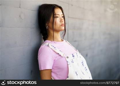 Young Chinese woman with a lost look and a serious expression leaning against the wall of a modern office building. Female wearing casual clothes.. Young Chinese woman with a lost look and a serious expression leaning against the wall of a modern office building.