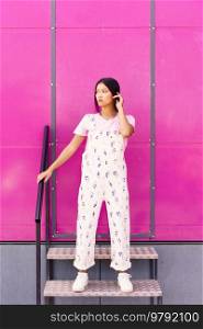 Young Chinese woman with blank stare and serious expression standing against pink wall of modern building. Asian Female in casual clothes.. Chinese woman with blank stare and serious expression standing against pink wall of modern building.