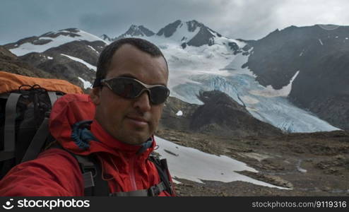 Young climber with red jacket and big backpack taking a selfie against mountains with glaciers in the background during a cloudy day. Young climber with red jacket and big backpack taking a selfie against mountains with glaciers in the background