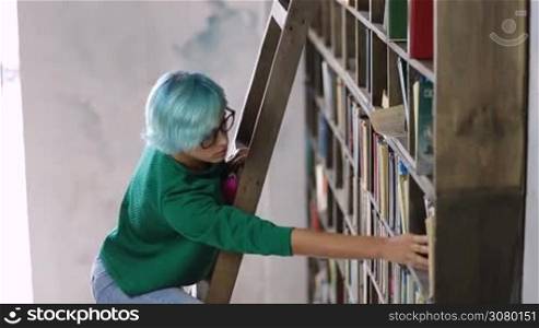 Young college female student in spectacles looking for a book on bookshelf in library while standing on wooden stepladder. Charming hipster woman with blue hair standing on ladder at bookshelf and picking the book.