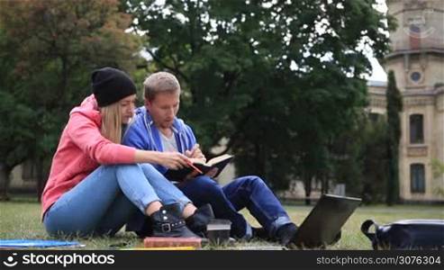 Young college friends sitting on campus lawn in the park and studying together. Couple of students reading a book and learning educational materials outside university.