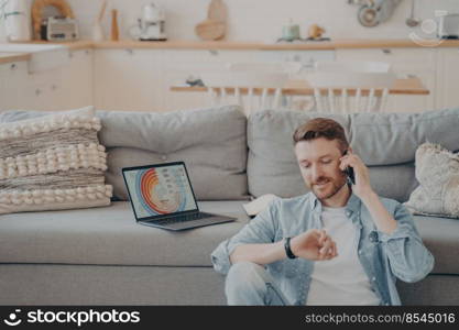 Young company male worker calling his supervisor while uploading infographics and sending them to his boss, checking time hoping he isn’t late, sitting on floor while resting against couch. Young company male worker calling his supervisor while sending infographics to boss