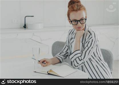 Young concentrated ginger woman in eyewear dressed casually thinking about new day while sitting at table in kitchen with transparent glass of water, looking aside while making notes in notebook. Young concentrated ginger woman thinking about new day while making notes in notebook at kitchen