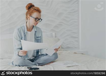 Young concentrated woman housewife in spectacles managing family budget while sitting on bed at home, redhead female calculating bills household expenses, holding papers with charts and graphics. Young concentrated woman housewife in spectacles managing family budget while sitting on bed at home