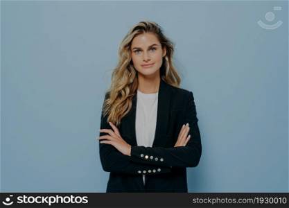 Young confident blonde business woman with long wavy hair dressed in smart black costume standing with crossed arms, posing over blue background in studio with copy space for advertising. Young confident blonde business woman dressed in smart black costume standing with crossed arms
