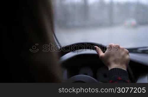 Young confident female driver holding steering wheel firmly with one hand while driving car on the freeway on winter day. Blurry view of inside car with bokeh during snow at daytime while woman driving car on highway.