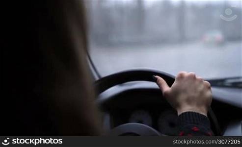 Young confident female driver holding steering wheel firmly with one hand while driving car on the freeway on winter day. Blurry view of inside car with bokeh during snow at daytime while woman driving car on highway.