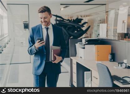 Young confident financier in formal classic suit with laptop and agenda, smiling and reading messages on smartphone, while walking down office corridor, rushing to company presentation meeting. Young confident financier in formal suit with laptop and agenda, reading messages on smartphone