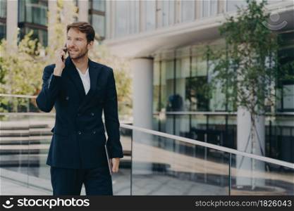 Young confident office worker in formal smart suit walking outside of business center, talking on mobile phone, holding laptop in his hand, hurrying to work on sunny morning. Young man office worker in suit talking on mobile phone while walking outside of business center