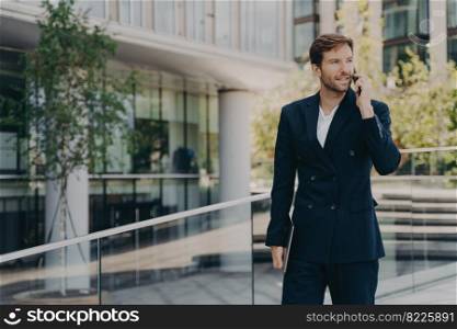 Young confident office worker in formal smart suit walking outside of business center, talking on mobile phone, holding laptop in his hand, hurrying to work on sunny morning. Young man office worker in suit talking on mobile phone while walking outside of business center
