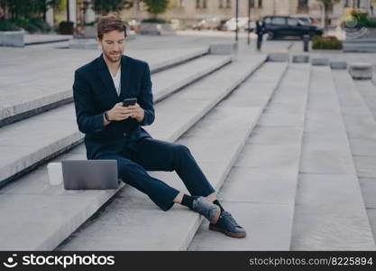 Young confident urban business professional man in stylish suit using smartphone while sitting on steps outside with laptop. Businessman holding mobile phone using app, typing messages or urgent email. Young confident man in stylish suit using smartphone while sitting on steps outside with laptop