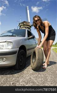 Young, confident woman, changing a flat tire on her car on a rural road with a wind mill in the backgrounc