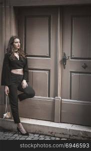 Young confident woman, in an elegant, modern black outfit, leaned against a wall, in front of a big vintage wooden door, looking up.