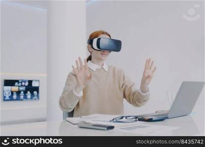 Young contemporary european woman in VR headset is working in office. Girl is sitting at the desk in front of computer and gesturing with hands. Concept of digital solutions and future innovations.. Young contemporary european woman in VR headset is working in office. Future innovations concept.