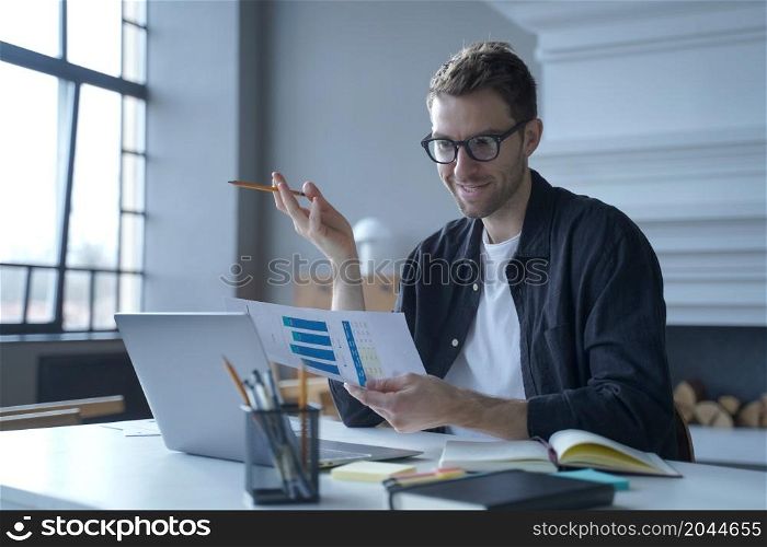 Young contented Austrian businessman in glasses discussing last deal transaction report with sales department via video call meeting on laptop computer while working remotely from modern home office. Contented Austrian businessman discussing last deal report with sales department via video call