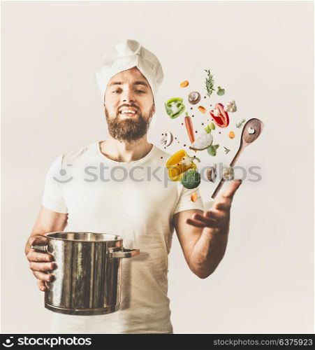Young cook man with hat and beard holding pan and spoon with flying vegetables on white background. Healthy cooking concept