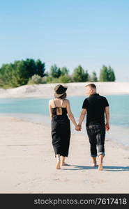 young couple a guy with a girl in black clothes are walking on the white sand at the edge of blue water