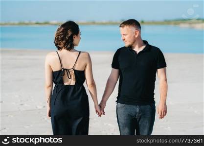 young couple a guy with a girl in black clothes are walking on the white sand at the edge of blue water