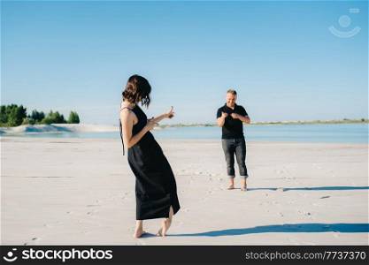 young couple a guy with a girl in black clothes are walking on the white sand at the edge of blue water