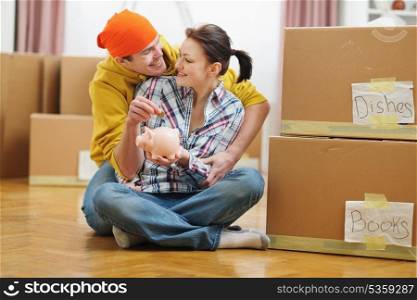 Young couple among boxes putting coin in piggy bank