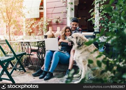 Young couple and their dog sitting smiling in the garden chairs near their wooden house with a laptop