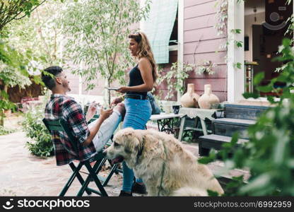 Young couple and their dog sitting smiling in the garden chairs near their wooden house