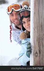 young couple at ski resort hiding behind wooden post