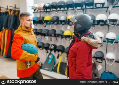 Young couple at the showcase trying on helmets for ski or snowboarding, side view, sports shop. Winter season extreme lifestyle, active leisure store, buyers looking on protect equipment