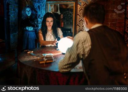 Young couple at the table with crystal ball on spiritual seance, scary witch reads the spell on background. Female foreteller calls the spirits. Couple on spiritual seance, witch on background