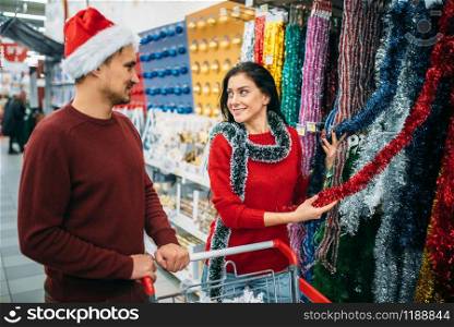 Young couple choosing holiday fluffy garland, purchase of christmas decoration in supermarket, family tradition. December shopping. Young couple choosing holiday fluffy garland