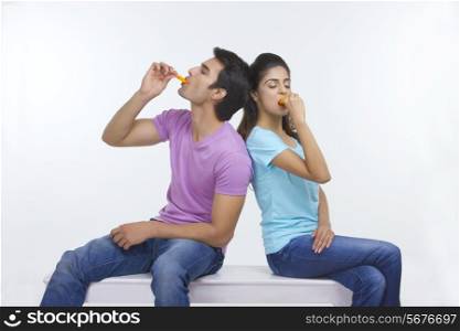 Young couple eating ice lollies while sitting on bench over white background
