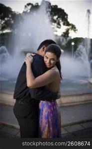 Young couple embracing by fountain in plaza, sunset