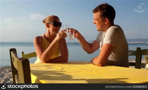 Young couple enjoying drinks at the seaside while on their summer vacation sitting at a table on the beach in the sunshine