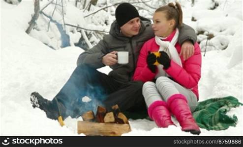 Young couple enjoying winter vacation near bonfire in snowy forest