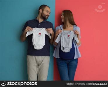 young couple holding baby bodysuits. Beautiful pregnant woman and her husband expecting baby holding baby bodysuits and smiling over colorful background