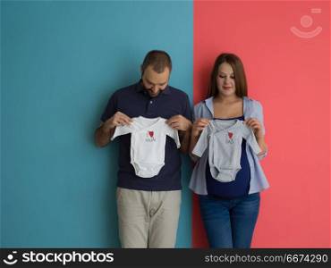 young couple holding baby bodysuits. Beautiful pregnant woman and her husband expecting baby holding baby bodysuits and smiling over colorful background
