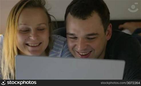 Young couple in bed spending evening with laptop. They watching humorous show or funny video in Internet