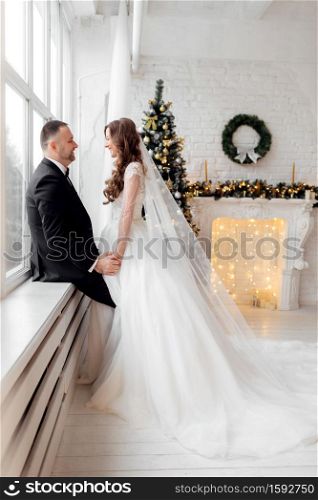 Young couple in love bride and groom posing in studio on background decorated with Christmas tree in their wedding day at Christmas. Enjoy a moment of happiness and love. Young couple in love bride and groom posing in studio on background decorated with Christmas tree in their wedding day at Christmas. Enjoy a moment of happiness and love.