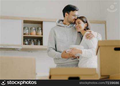 Young couple in love embrace and kiss with tender, hold pile of white plates, stand in kitchen during moving day, surrounded with many carton boxes filled with personal belongings, unpack stuff