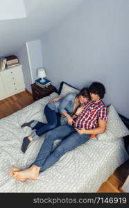 Young couple in love embraces looking electronic tablet while resting over a bed. Leisure time at home concept.. Couple in love looking electronic tablet on bed