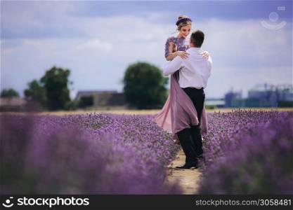 Young couple in love hugging and walking in a lavender field on summer cloudy day. girl in a luxurious purple dress and with hairstyle.. Young couple in love hugging and walking in a lavender field on summer cloudy day. girl in a luxurious purple dress and with hairstyle