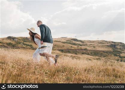 Young couple in love on the mountain range in summer or autumn day walking in nature freedom she is holding his hand and leading wearing hat side view